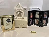 Photo showing 5 assorted picture frames: gold-tone rectangular, round white Lenox frame on box, black folding multi-photo frame, and two white Lenox boxes in background.