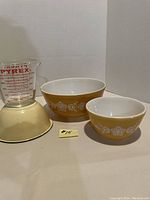 Set of two yellow Pyrex floral patterned mixing bowls, enamel bowl, and a Pyrex 2-cup measuring cup with red measurement markings shown in front of white background.