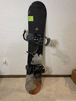 Front view of snowboard standing upright showing black surface with bindings installed.