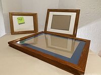 Photo of 3 wooden wall frames on the floor against a white wall. One non-matted 7.5" x 9.5" frame on the left, one matted 8.5" x 7.5" with white mat in middle, and one matted 16" x 12" with royal blue mat on bottom.