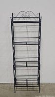 Front view of black wrought iron baker's rack with 4 slatted shelves and scrollwork detail.