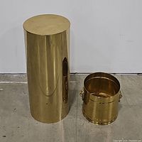 Brass pedestal column and brass planter/trash can side by side on a concrete floor.