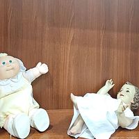 Two dolls side by side on wooden shelf showing Baby Jesus porcelain doll in white gown and vintage yellow clad Cabbage Patch doll.