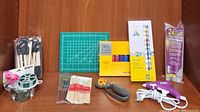 All crafting items displayed on a wooden surface including two acrylic paint boxes, paint brushes in plastic packaging, rotary cutter, cutting mat, glue gun, floral wire, foam brushes, wooden paint sticks, and other accessories.
