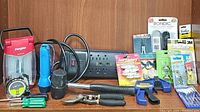 Photo showing various garage tools and hardware on a wooden surface including flashlight, screwdrivers, surge protector, pliers, measuring tape, and small tools.