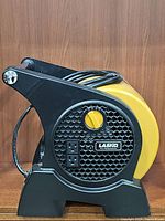 Front angle view showing the black honeycomb grille, yellow speed control dial, two power outlets, and Lasko badge on the side of the yellow fan body.