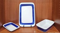 Photo showing all three items: two Koka platters by Hertha Bengtson for Rörstrand Sweden with blue leaf patterned borders, and one rectangular casserole dish with a solid blue band on white ceramic.
