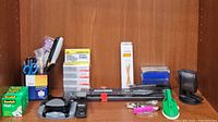 Photo showing full assortment of office supplies including tape dispensers, staplers, staples boxes, scissors, pencils, hole puncher, and additional miscellaneous items on wooden shelf.