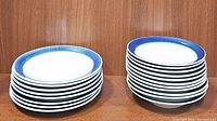 Stack of 8 dinner plates with a stack of 12 matching bowls beside them, displaying white ceramic with blue rims.