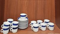 12 white china cups with blue textured bands around the rim, each with a handle. 12 matching saucers stacked beside them.