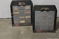 Two black Rubbermaid plastic compartment storage boxes with multiple drawers, placed on the floor.