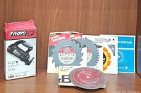 RotoZip spiral saw in its box alongside five different packaged saw blades of various types and sizes and one loose blade in front.