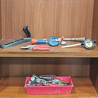 Tools arranged on wooden shelf: metal hand plane, red wire shears, two screwdrivers with blue and orange handles, vintage hand drill, Flotec drill pump, and pink tray with assorted small tools.