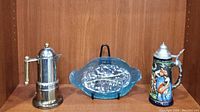 Wide shot showing all three items together on shelf: King beer stein, blue dish on stand, and metal stovetop espresso maker with brass handle