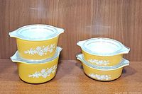Four stacked yellow Pyrex casserole dishes with butterfly gold floral patterns and clear glass lids, arranged on a wooden surface.