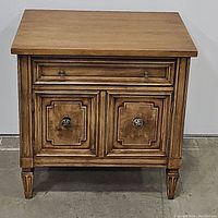 Front view of vintage wooden nightstand showing one drawer and two doors with detailed wood paneling and metal knobs.