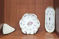 Three porcelain plates displayed on wood shelf, including oval tray with gold and black detailing, floral shaped plate with gold trim, and white textured triangular dish