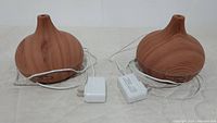 Two wood grain aroma diffusers with attached power cords and adapters on beige fabric surface, showing overall items.