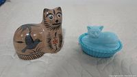 Two cat figurines shown together on a white cloth background, a tan colored Mexican Tonala pottery cat with hand-painted birds and floral designs next to a solid blue Westmoreland milk glass cat on a basket base.