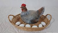 Top-down and angled views of the ceramic chicken sitting in the woven basket surrounded by seven ceramic eggs, displayed on a white quilted surface.