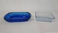 Shows the top view of the oblong cobalt blue Anchor Hocking dish and a square blue tinted dish placed side by side on a white quilted surface.