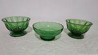 Three emerald green glass bowls displayed side by side on white surface. Two pedestal bowls on the left and right sides and one scalloped edge bowl in the center.