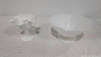 Two pieces of milk glass on a quilted surface. Left is a Fenton Hobnail compote with ruffled edge and pedestal, right is a smooth footed bowl by Anchor Hocking with octagonal shape.