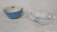 Two blue glass mixing bowls with white interior and floral pattern next to a white CorningWare baking dish with blue floral design and clear glass lid