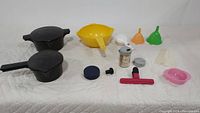 Wide shot of all plastic kitchen gadgets showing two strainer pots, funnels, bowls, measuring tool, and clips arranged on a white quilted surface.