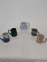 Shows the entire lot of one pitcher and four mugs arranged on a white quilted surface.
