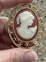 Hand-held close-up image of the cameo pendant showing white female profile with flower and gold-tone ornate frame.