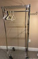 Metal clothes hanging rack with shelves and horizontal hanging bar, on rollers, shown against beige wall.