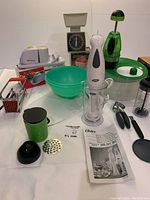Kitchen lot showing a variety of tools including the green French fry slicer with black plunger and slicing disc, an Oster stick mixer with mixing cup, user manual, a green mixing bowl, small kitchen scale, metal tongs, and additional slicer tool.