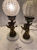 Pair of cherub lamps with glass shades and marble bases on white surface.