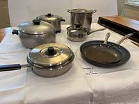 Collection of the cookware pots and fry pans displayed on a white cloth background with lids and handles visible