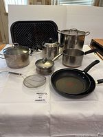 Wide shot of all cookware items including pots, pans, grill pan, kettle and strainer arranged on white table