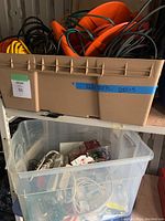 Plastic storage bins with coiled outdoor electrical cords in top bin, assorted indoor cords and power strips in clear bin below. Bins organized on a shelf in a garage.