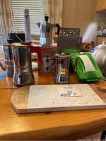 Espresso makers, napkin holder, crumb brush, green tape dispenser, marble paper towel holder, stone cutting board, and stainless steel mugs arranged on table.