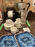 Espresso maker, coffee grinder, espresso cups with saucers, and kitchen hot pads arranged on kitchen counter.