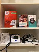 Photo showing iLife Blood Pressure Monitor box and device with cuff, and DirectAlert Mobile GPS+ box and owner's manual on shelf.