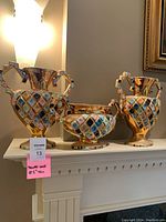 Set of three gold-colored decorative Italian vases with multicolored diamond pattern, displayed on mantlepiece under soft lighting.