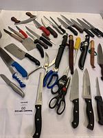 A large assortment of knives, cleavers, kitchen shears, sharpening steel and a curved meat cutter arranged on a white surface.