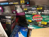 Assorted lightbulbs including Philips ColorTone outdoor flood bulbs in green and red, 65W replacement energy-saving bulbs 4 pack, and other packaged bulbs.
