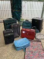 Group of multiple luggage pieces: hard and soft shell suitcases, garment bag, and travel bags in various colors including blue, black, red, green, and patterned design.