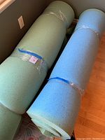 Two rolled rolls of memory foam mattress pads, one green and one blue, wrapped in plastic.