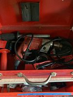 Opened red metal carrying case showing Milwaukee red drill and accessories inside including cable and drill handle.