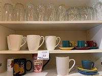 Shelf with tall clear arc glasses, white embossed mugs, colorful small espresso cups on saucers, and additional assorted mugs.