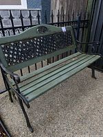 Full side view of the green painted wooden and black wrought iron bench outdoors, showing seat slats, armrests, and backrest lattice.