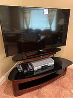 Full view of the Samsung flat screen TV placed on wood and glass TV stand, VHS and DVD players and multiple remote controls on the stand.