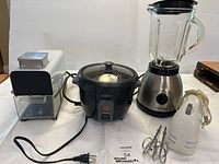 Four small kitchen appliances on a kitchen counter including a parmesan cheese grater, rice cooker, blender, and hand mixer